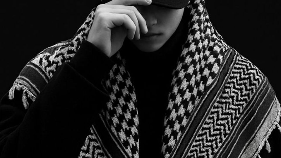 Studio portrait against a plain dark background, with minimal visible environment. The frame is tightly cropped, showing a black cap and a patterned fringed shawl dominating the foreground.