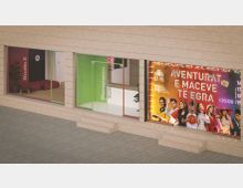 Street-level view of a modern building facade with large glass storefront windows and color-accented frames, including entrances labeled “Studio 2” and “Renaissance.” A brightly lit event poster fills one window, and shallow steps lead up from a paved sidewalk.