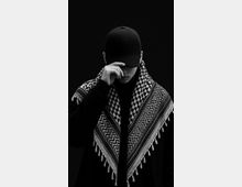 Studio portrait against a plain dark background, with minimal visible environment. The frame is tightly cropped, showing a black cap and a patterned fringed shawl dominating the foreground.